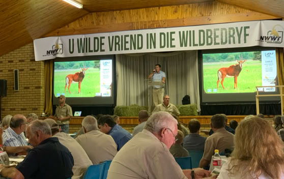 A group of people seated at tables in a hall with a wooden ceiling, watching a presentation. Two screens display an image of an animal, and a person is speaking on stage. A banner above reads "U Wilde Vriend in die Wildbedryf."